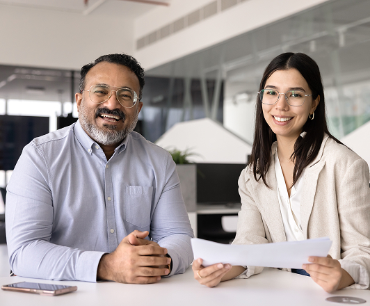 Two professionals sitting at a table in an office, smiling and looking at the camera, with laptops and papers in the background.